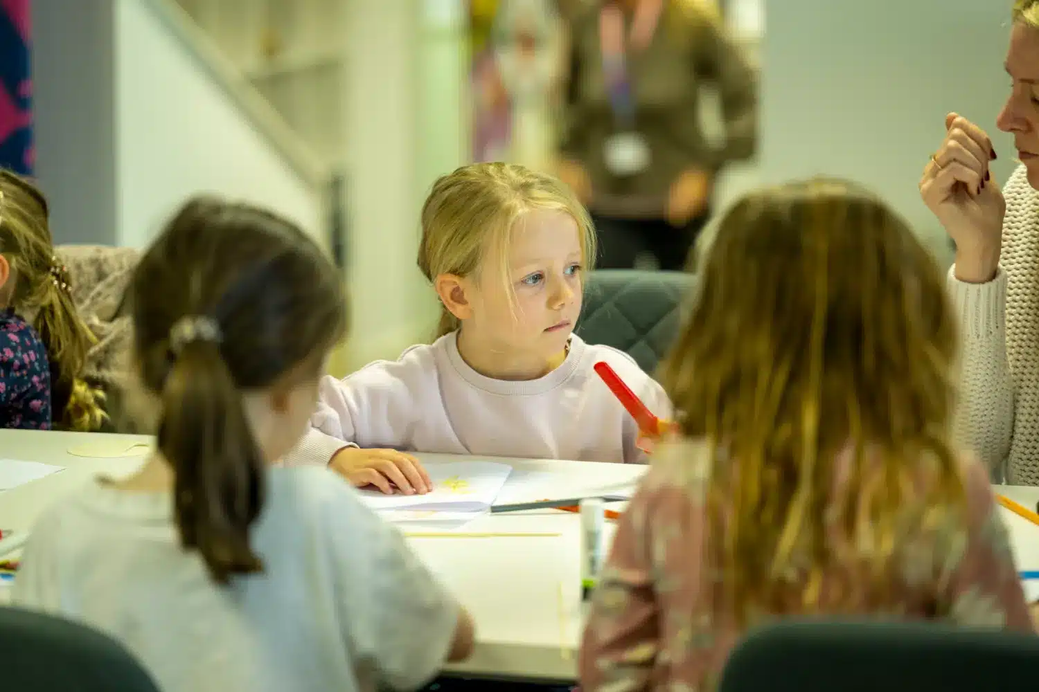 Three young girls sit at a table drawing with coloured markers and paper. An adult sits nearby, partially visible. The background is blurred, showing more people and a staircase. The focus is on the children.