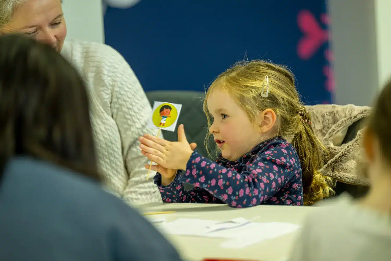 A young girl holds a paper puppet and talks to an adult woman at a table, with other people blurred in the foreground.