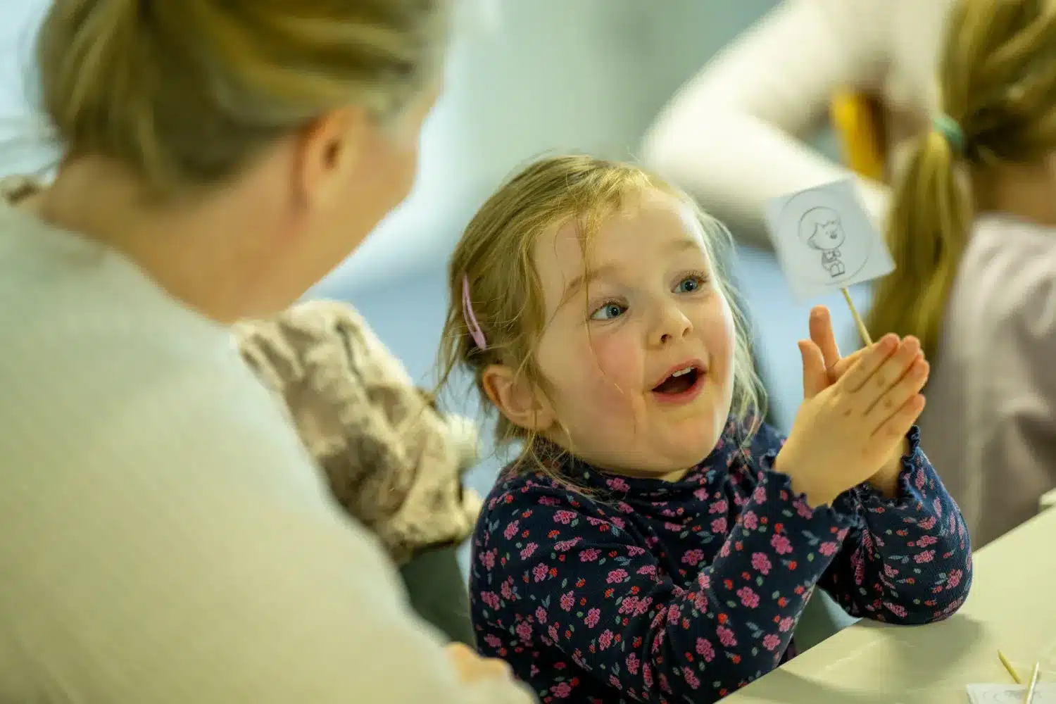 A young girl in a floral jumper excitedly holds up a small drawing on a stick, sitting at a table next to an adult with blond hair during a creative activity.