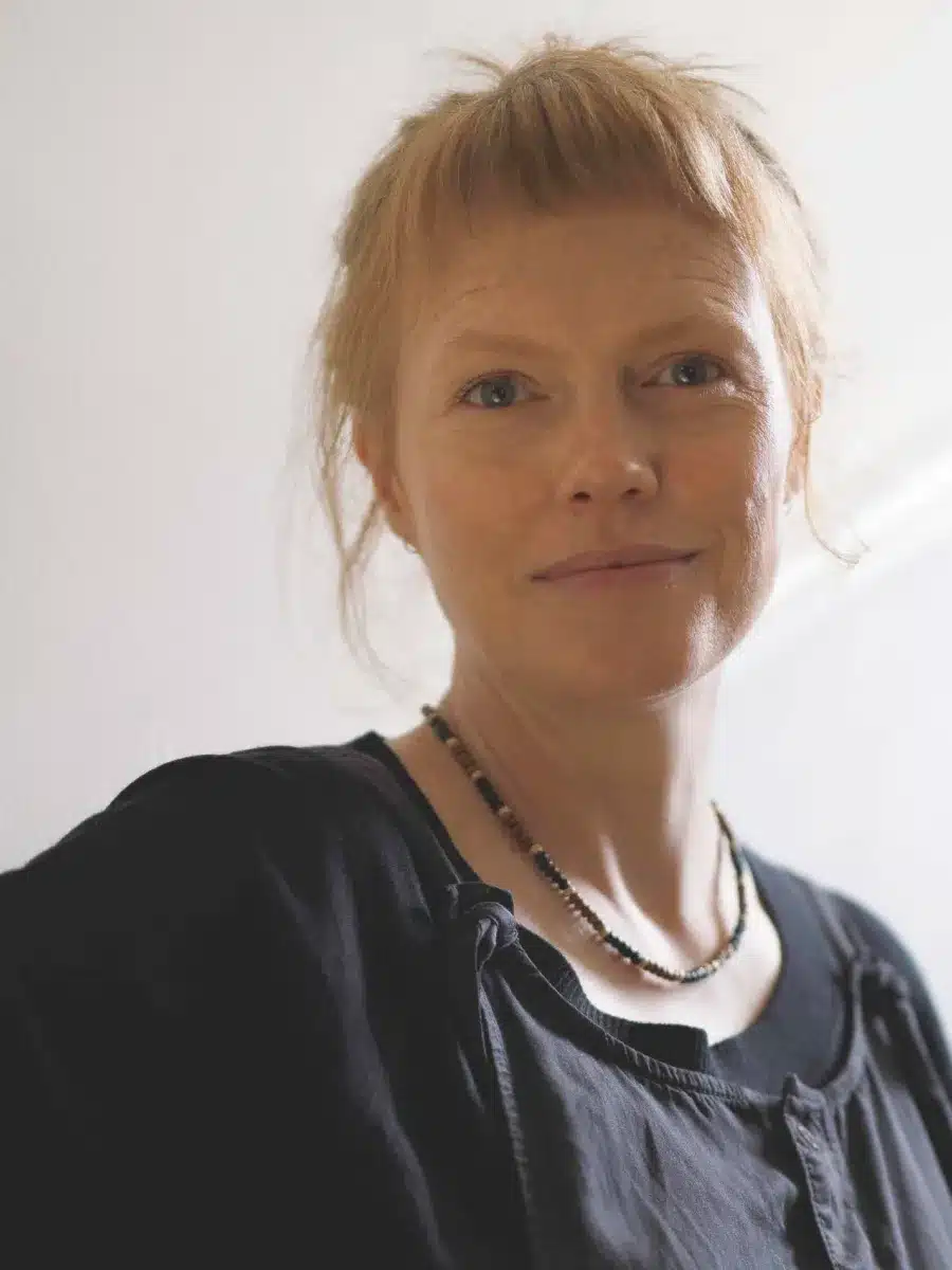 A woman with light skin and reddish-blonde hair pulled back, wearing a black top and necklace, stands against a light-coloured wall, softly smiling at the camera.