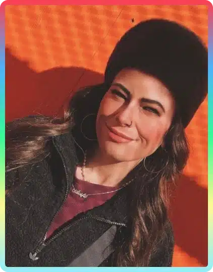 A woman with long brown hair, wearing a black fuzzy hat and jacket, smiles while squinting in bright sunlight against an orange wall at the Kilkenny Animated Festival. She has hoop earrings and a necklace.