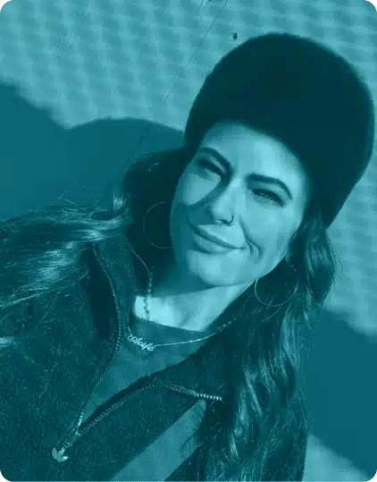 A person with long hair, wearing a furry hat, hoop earrings, and a zip-up jacket, smiles whilst standing outdoors in bright sunlight.