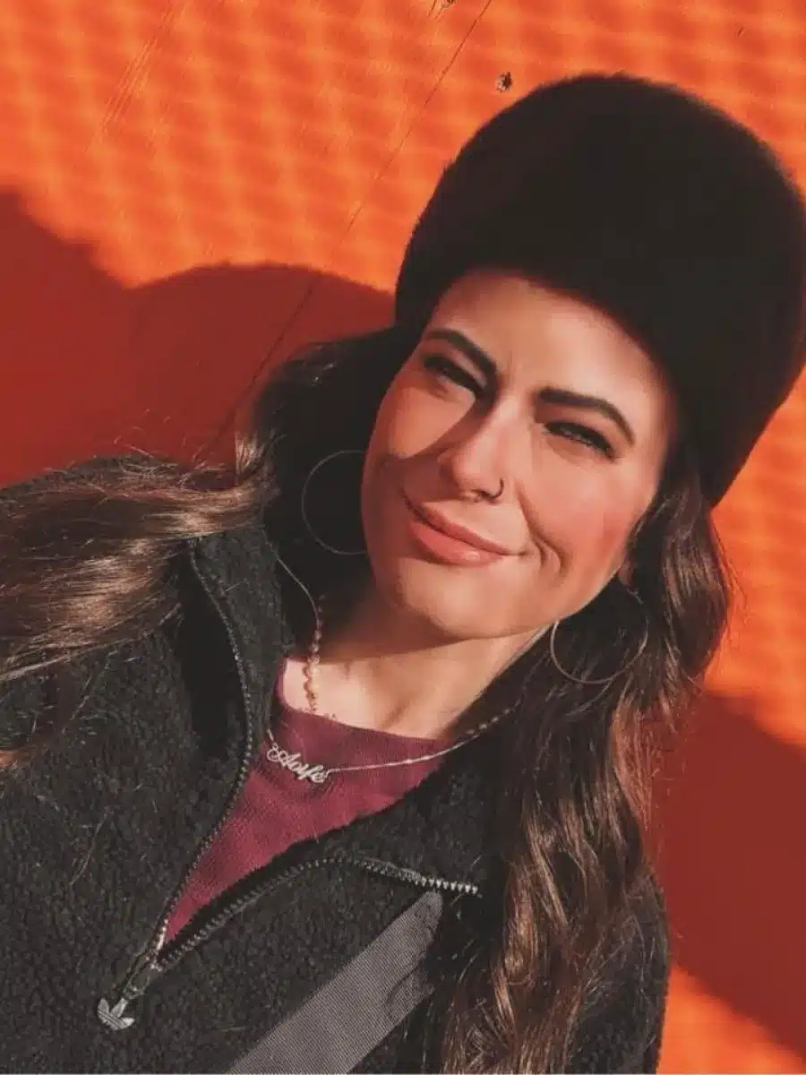A woman with long brown hair, wearing a black furry hat, hoop earrings, and a black jacket, stands against an orange textured background, smiling slightly.