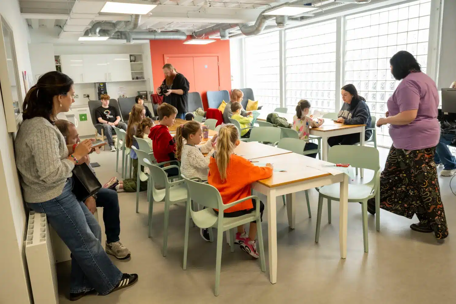 Children sit at tables in a classroom, working on assignments. Two adults stand nearby, one writing on a clipboard and another engaging with pupils. A photographer is also present in the background.