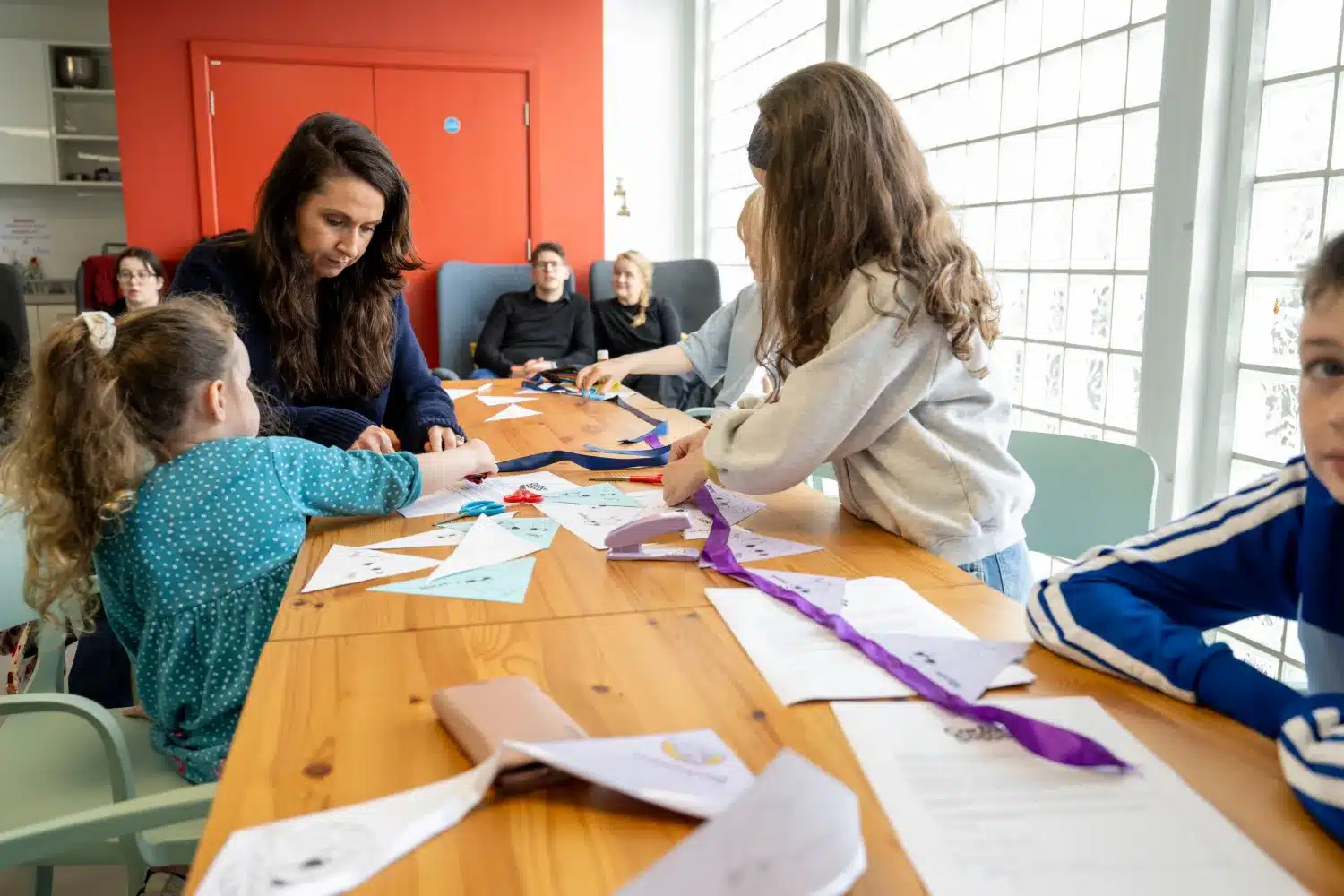 Children and an adult sit at a wooden table, making crafts with paper and ribbons in a bright room with large windows. Papers, colourful materials, and instructions are spread across the table. Other people sit in the background.