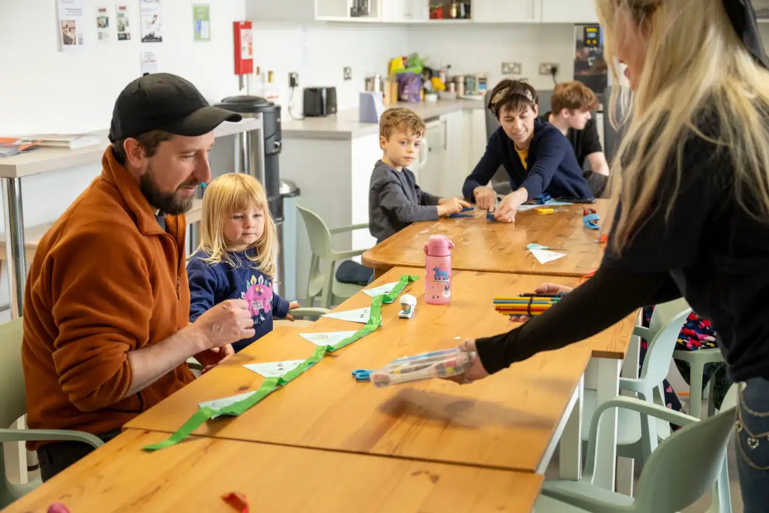 Adults and children sit at a table doing arts and crafts. A woman hands supplies to a girl and a man. Felt-tip pens, paper, and decorations are spread out on the table. The setting is a bright room with a kitchen area in the background.