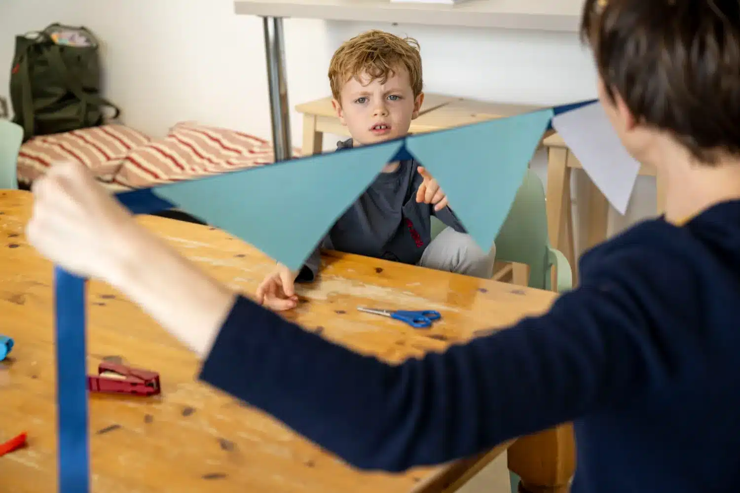 A child sits at a wooden table watching an adult hold a string of triangular blue flags. Scissors and crafting supplies are on the table, suggesting they are working on an arts and crafts project together.