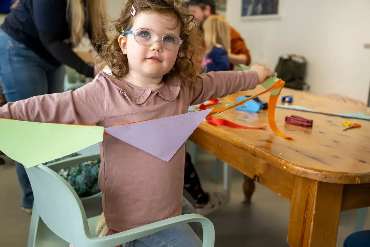 A young girl with glasses sits on a chair, holding a colourful paper bunting. She is in a craft room with other children, a wooden table with art supplies, and adults in the background.