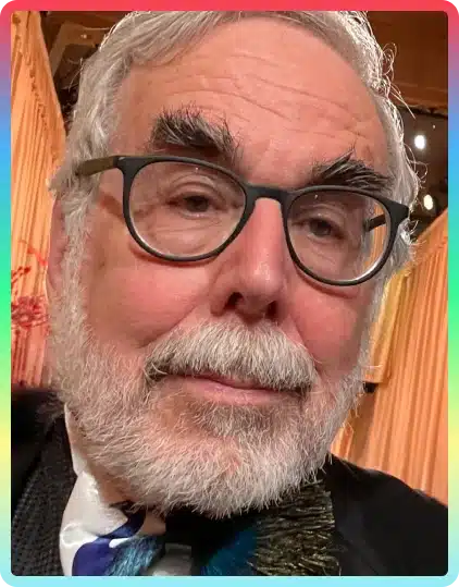 An older man with grey hair, thick eyebrows, glasses, and a white beard is wearing a suit and sitting indoors with warm lighting and curtains in the background, perhaps ready to share his insights at the Kilkenny Animated Festival.