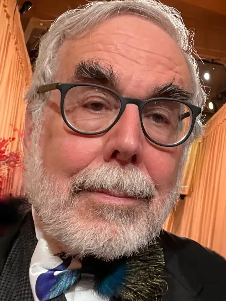 An older man with grey hair, thick eyebrows, and a white beard wears glasses, a dinner jacket, and a multicoloured bow tie, posing for a selfie at an indoor event with soft lighting and tan curtains.