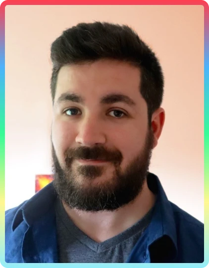 A man with dark hair and a full beard is smiling softly. He wears a blue shirt over a grey T-shirt. The background is plain, with soft pink tones, and the photo—framed by a colourful border—captures the cheerful spirit of the Kilkenny Animated Festival.