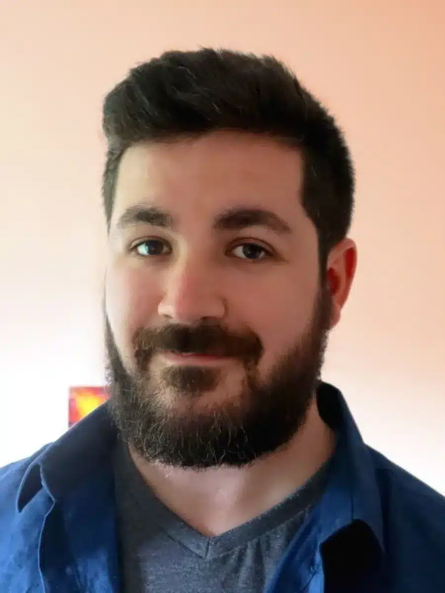 A man with dark hair and a full beard smiles slightly at the camera. He is wearing a blue shirt over a grey T-shirt, standing in front of a plain, light-coloured wall.