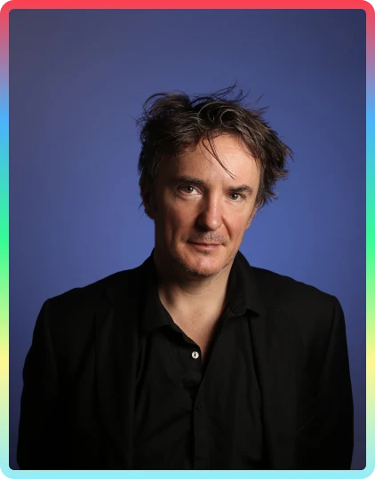 A man with tousled dark hair wearing a black shirt and blazer poses in front of a blue background, framed by a colourful gradient border, evoking the vibrant spirit of the Kilkenny Animated Festival.