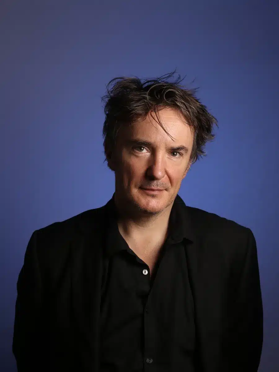 A man with tousled dark hair wearing a black shirt and jacket stands against a solid blue background, looking directly at the camera with a neutral expression.
