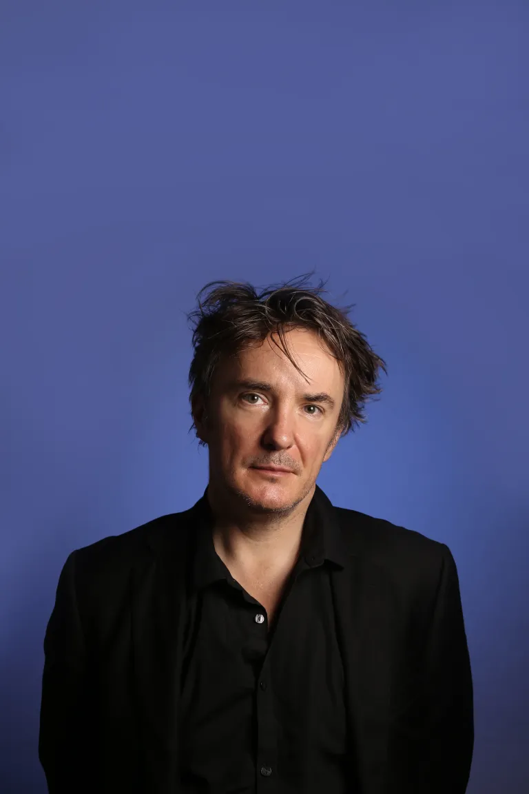 A man with tousled dark hair wearing a black shirt and jacket stands against a plain blue background, looking slightly to the side with a neutral expression.