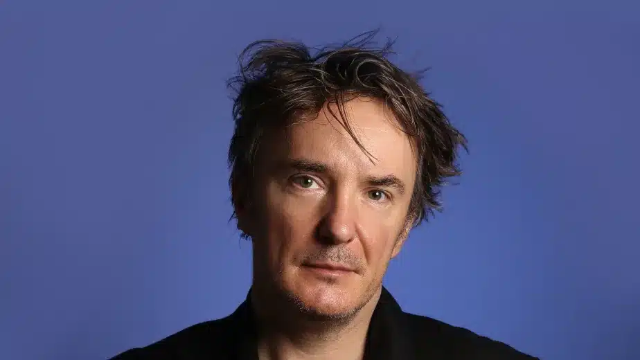 A man with tousled dark hair wearing a black shirt and jacket stands against a plain blue background, looking slightly to the side with a neutral expression.
