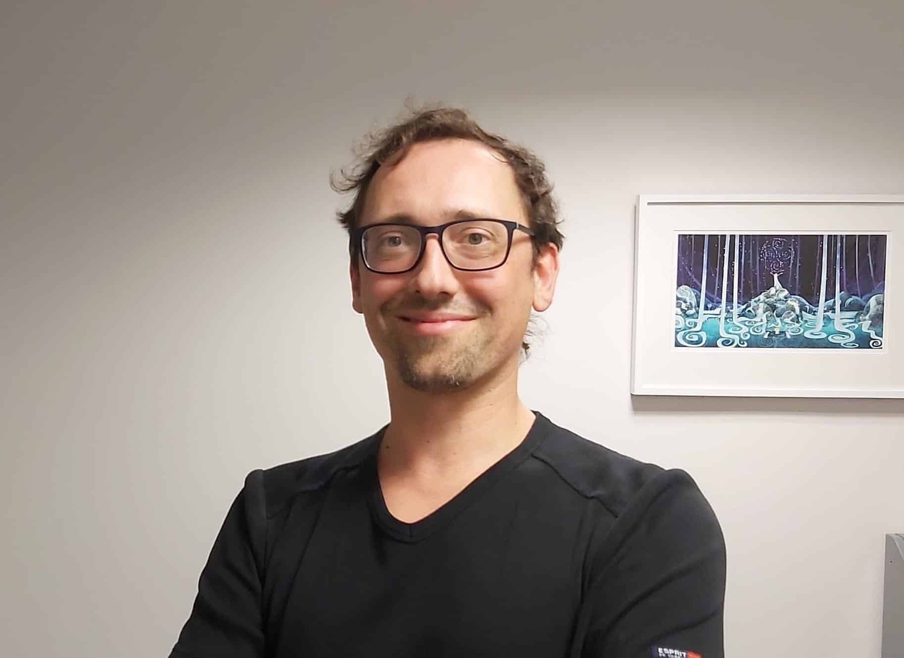 A man with glasses and short brown hair smiles whilst standing with arms crossed. He wears a black shirt and stands in front of a white wall with a framed artwork showing a mystical forest scene.