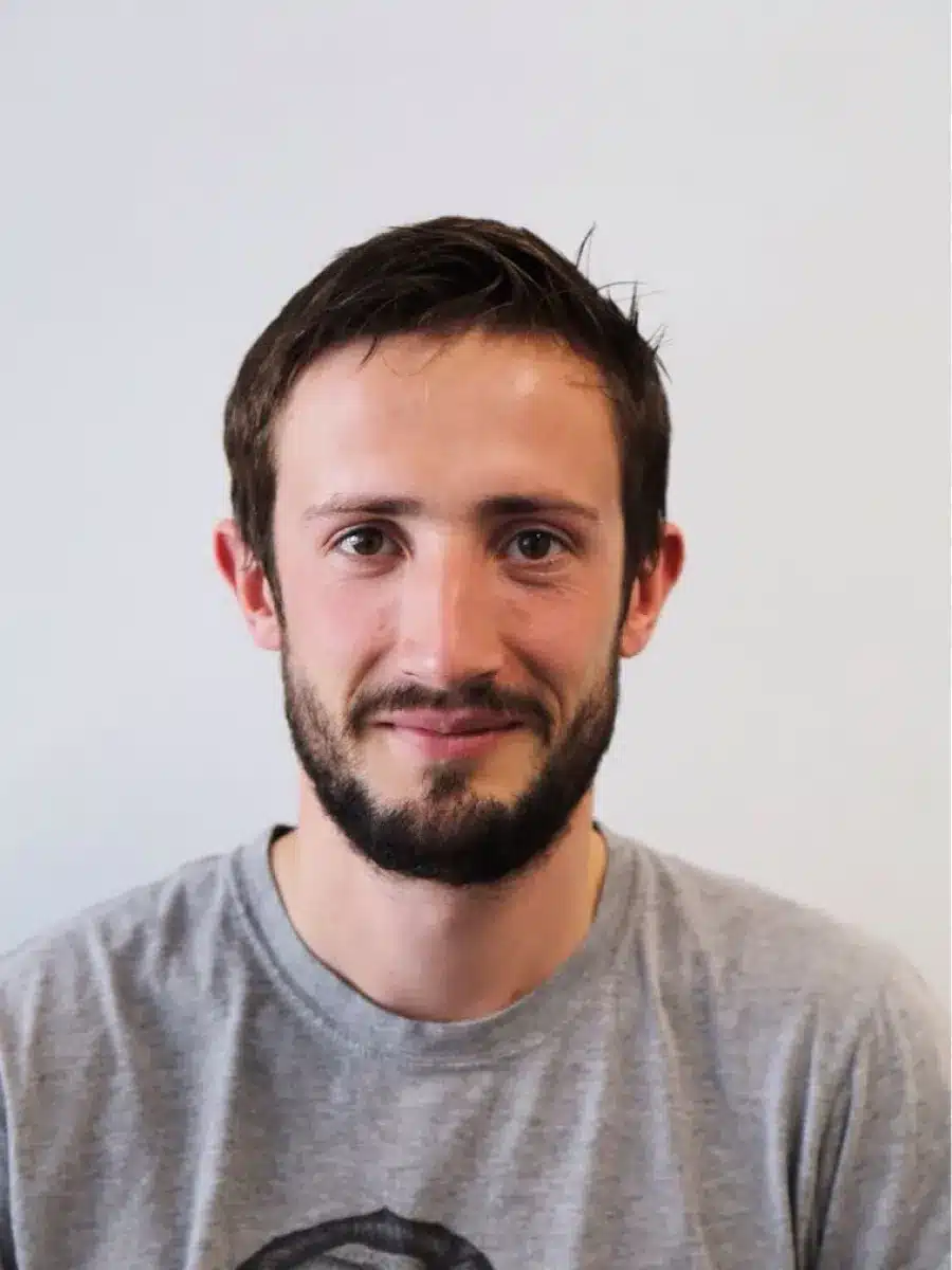 A young man with short brown hair and a beard, wearing a light grey t-shirt, smiles softly at the camera against a plain light background.