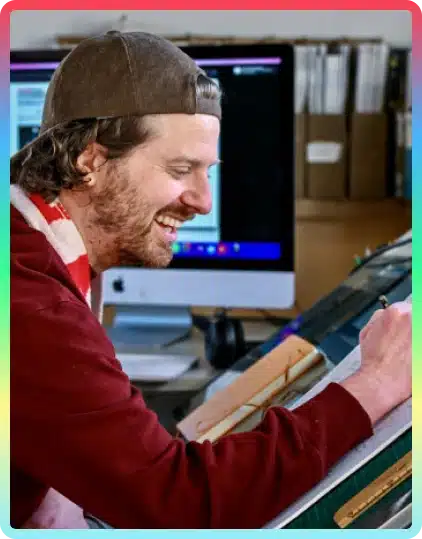 A smiling person wearing a backwards cap and red shirt draws on paper at a desk, perhaps inspired by the creativity of Kilkenny Animated Festival, with a computer and shelves in the background.