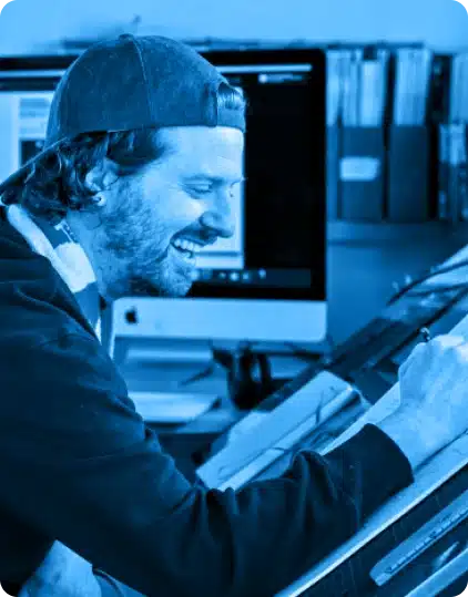 A smiling man wearing a backwards cap draws at a desk with a computer and bookshelves in the background. The image, with its blue tint, captures the creative atmosphere often found at events like the Kilkenny Animated Festival.