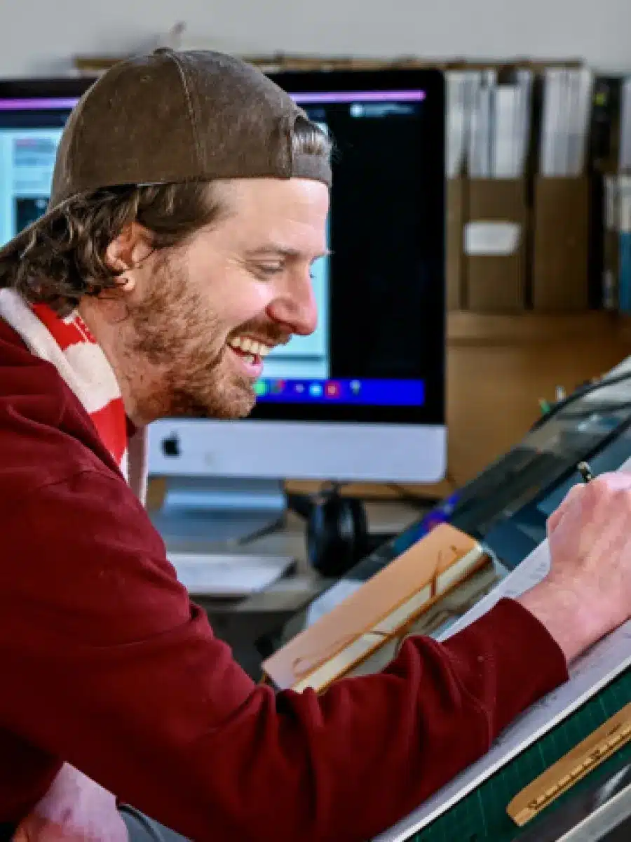 A man in a red jumper and backwards cap smiles while drawing at a drawing table, with computer monitors and bookshelves in the background.