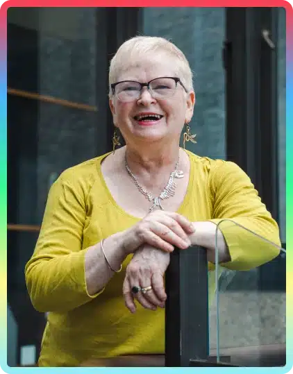 Smiling older woman with short white hair, glasses, and a yellow top stands outdoors, leaning on a railing. She wears statement earrings and a necklace, with a rainbow-coloured border framing the photo—capturing the cheerful spirit often seen at the Kilkenny Animated Festival.