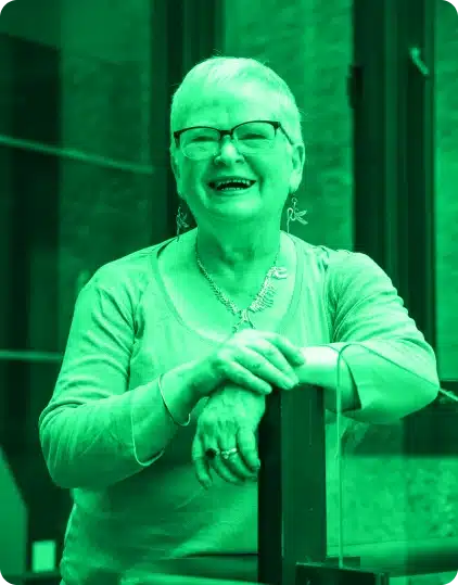 An older woman with short hair and glasses smiles whilst leaning on a glass surface, exuding the creative energy of the Kilkenny Animated Festival. She wears a necklace, earrings, and a long-sleeve shirt. The image has a green filter.