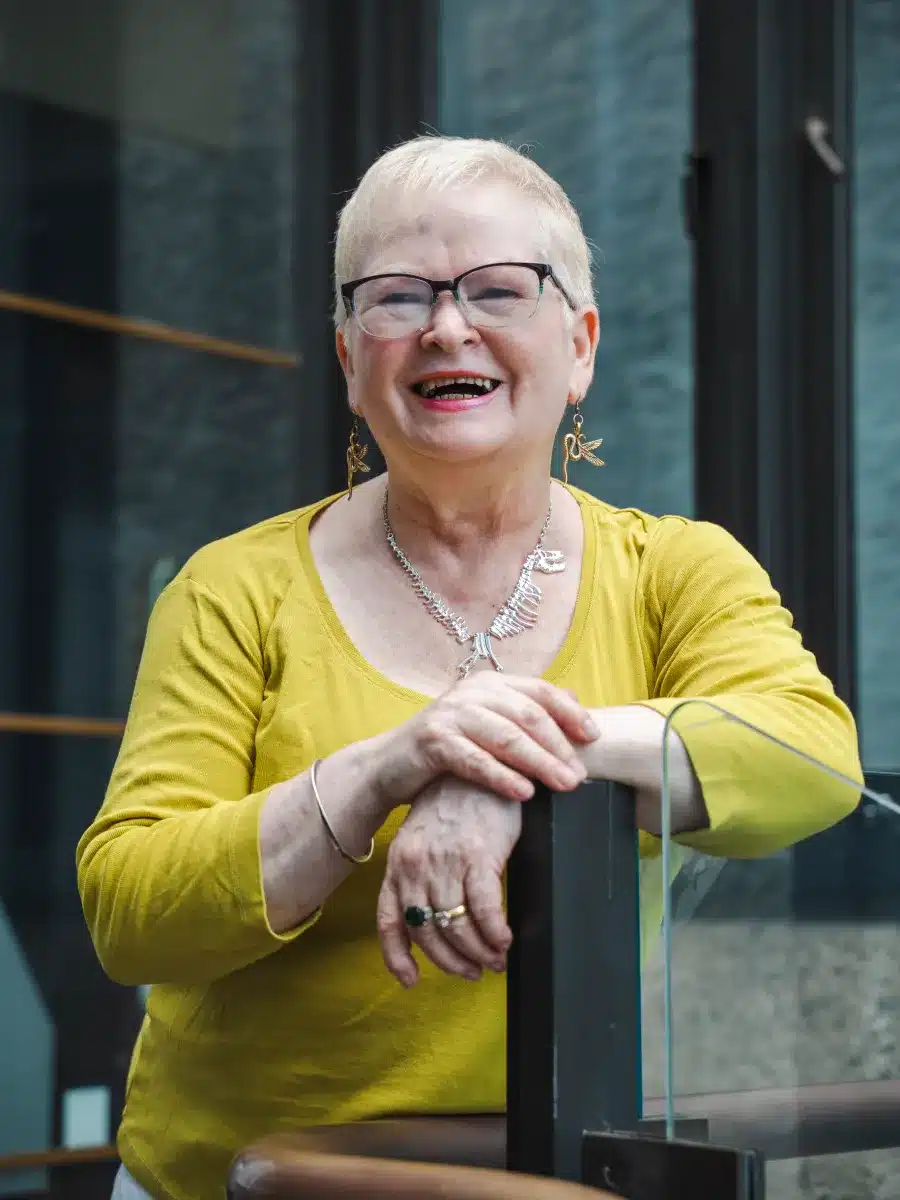 An older woman with short, light hair and glasses smiles warmly, leaning on a railing indoors. She wears a yellow top, silver jewellery, and earrings, standing in front of a modern glass and stone background.