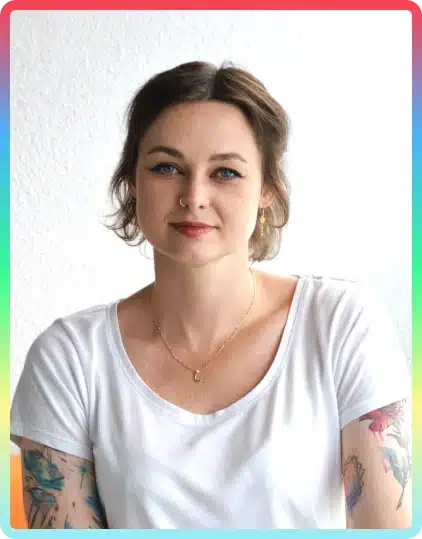 Woman with short brown hair and tattoos on her arms wears a white shirt and gold necklace, sitting against a white background with a colourful border around the photo—perfectly capturing the creative spirit of the Kilkenny Animated Festival.