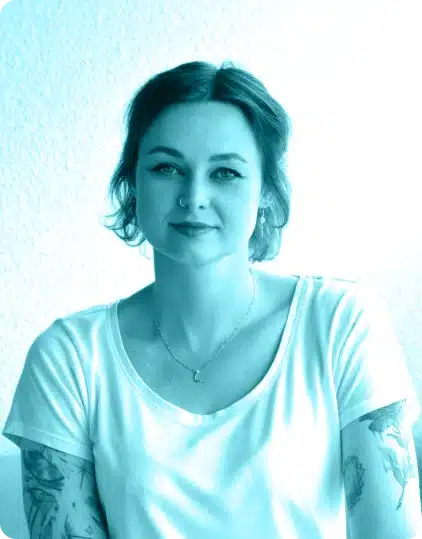 A woman with short hair and tattoos on her arms, wearing a white T-shirt and necklace, sits against a light-coloured background, looking at the camera with a slight smile. The image, reminiscent of portraits from the Kilkenny Animated Festival, has a blue tint.
