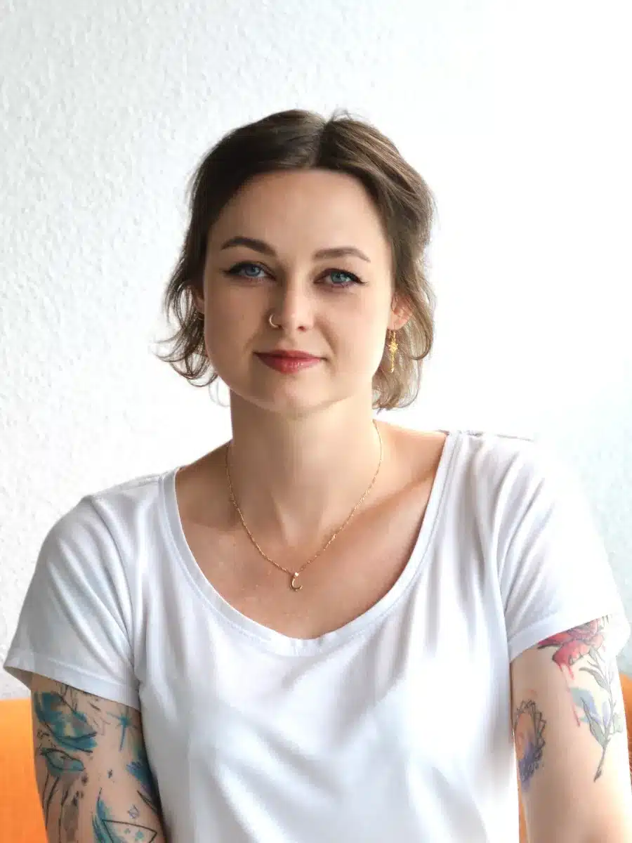 A woman with brown hair, blue eyes, and tattoos on her arms sits against a white background. She is wearing a white shirt, gold jewellery, and is smiling softly at the camera.
