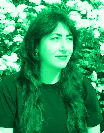 Person with long wavy hair wearing a dark shirt sits in front of blooming flowers at the Kilkenny Animated Festival. The entire image has a bright green filter.
