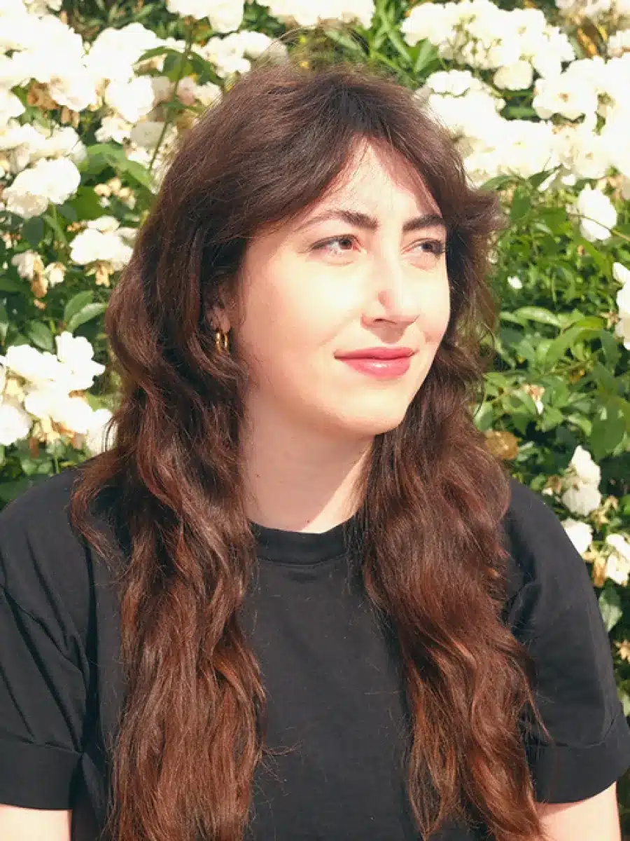 A person with long brown hair wearing a black shirt sits outdoors in front of blooming white flowers, looking slightly to the left with a neutral expression.
