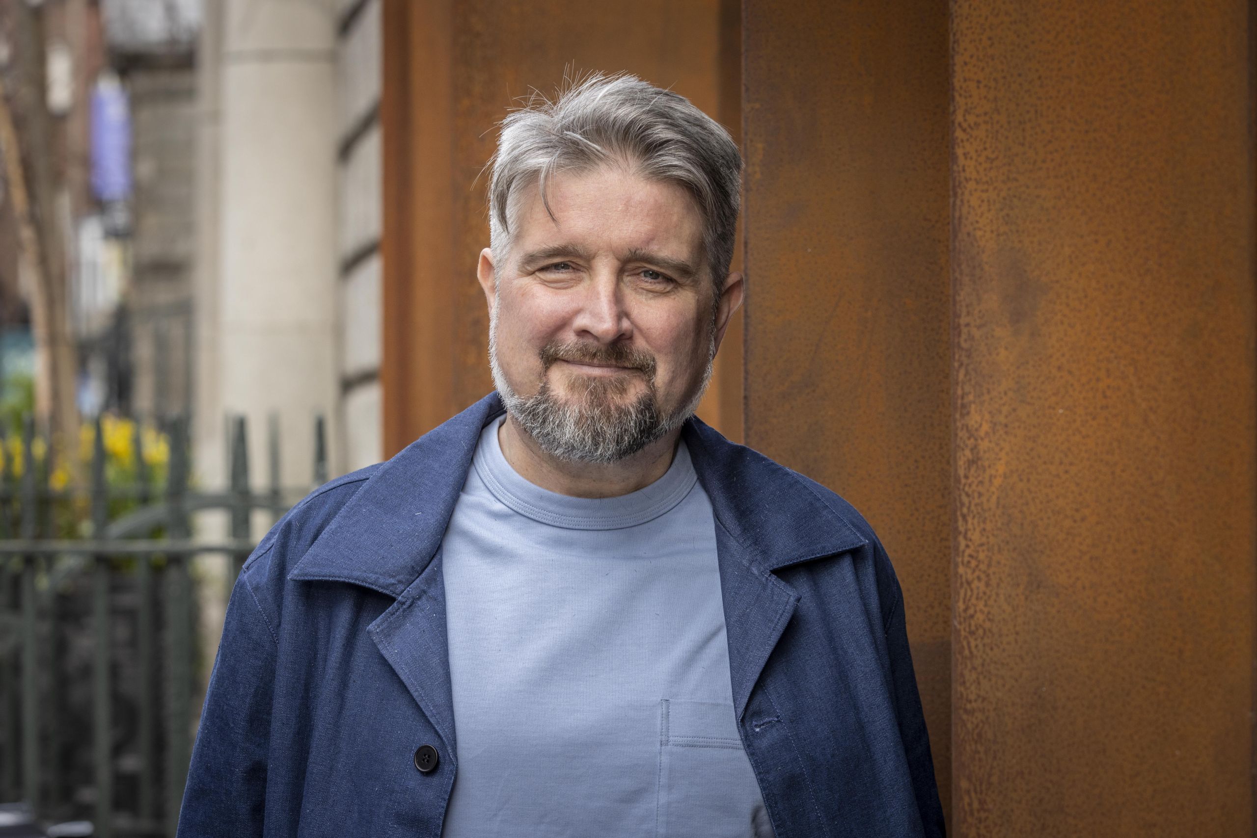 Middle-aged man with grey hair and beard, wearing a light blue shirt and dark blue jacket, stands outdoors near a rust-coloured wall and wrought-iron fence, smiling at the camera.