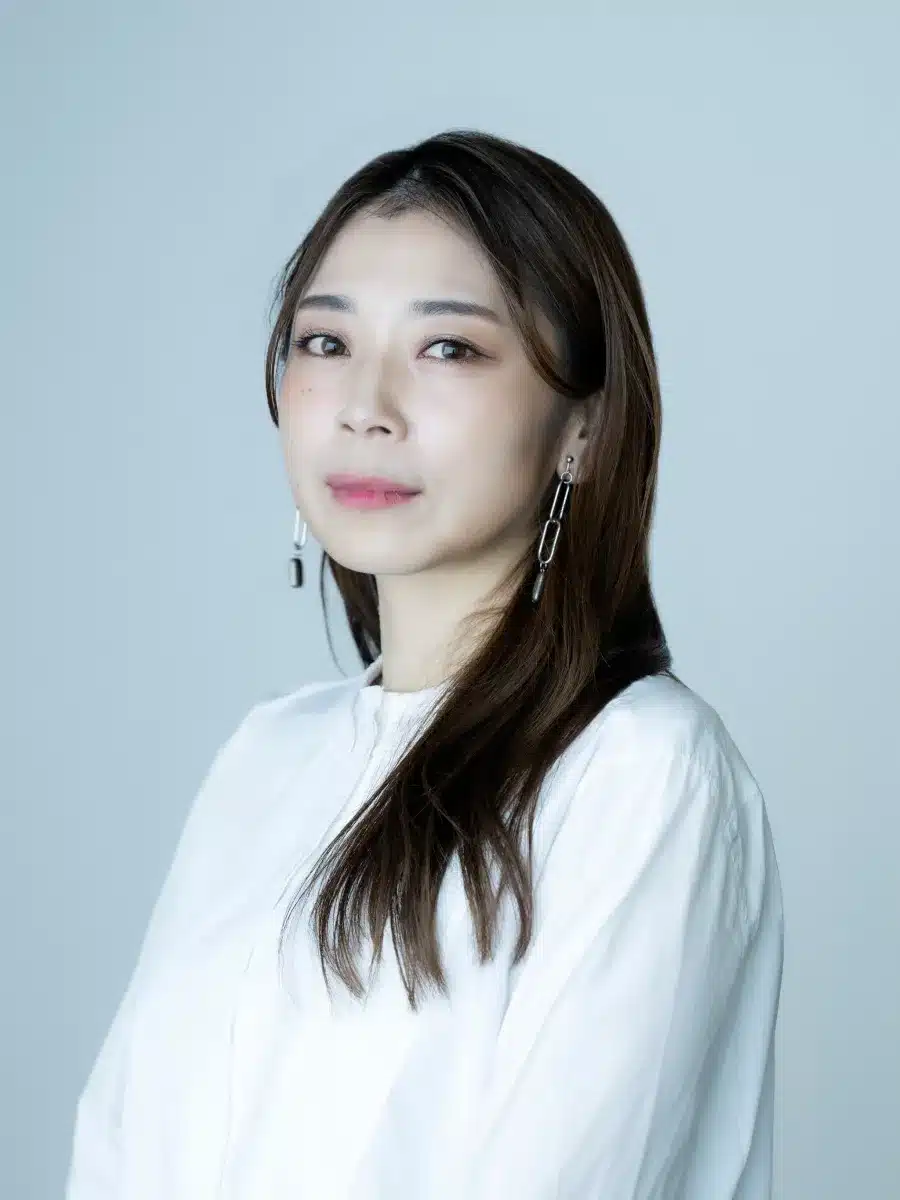 Woman with long brown hair wearing a white blouse and dangling earrings, standing against a plain light grey background, looking slightly to the side with a neutral expression.