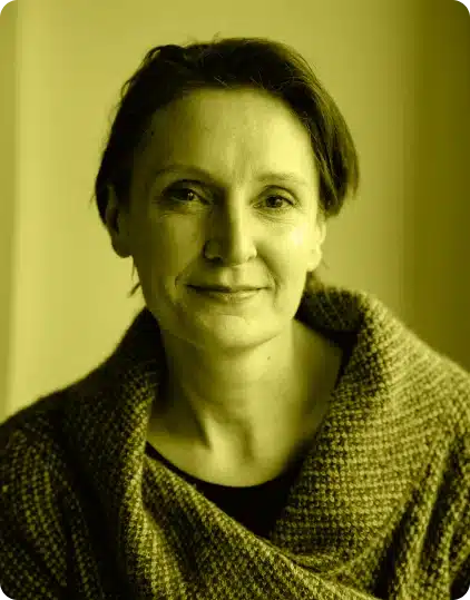 A woman with short hair, wearing a textured knitted jumper, looks at the camera with a gentle smile. The image has a yellowish tint and soft lighting, evoking the warm and inviting atmosphere of the Kilkenny Animated Festival.