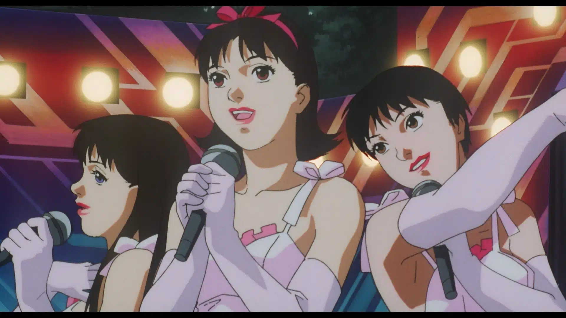 Three animated female singers in matching pink and white outfits and gloves perform on stage under colourful lights, each holding a microphone and smiling.