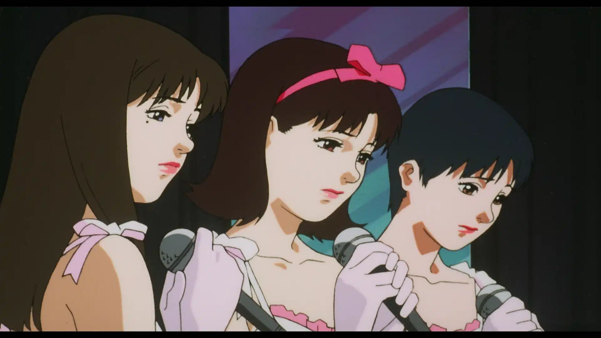 Three animated young women in pink outfits and gloves hold microphones, standing close together on stage, appearing serious or thoughtful, with a dark background and a colourful reflective surface behind them.