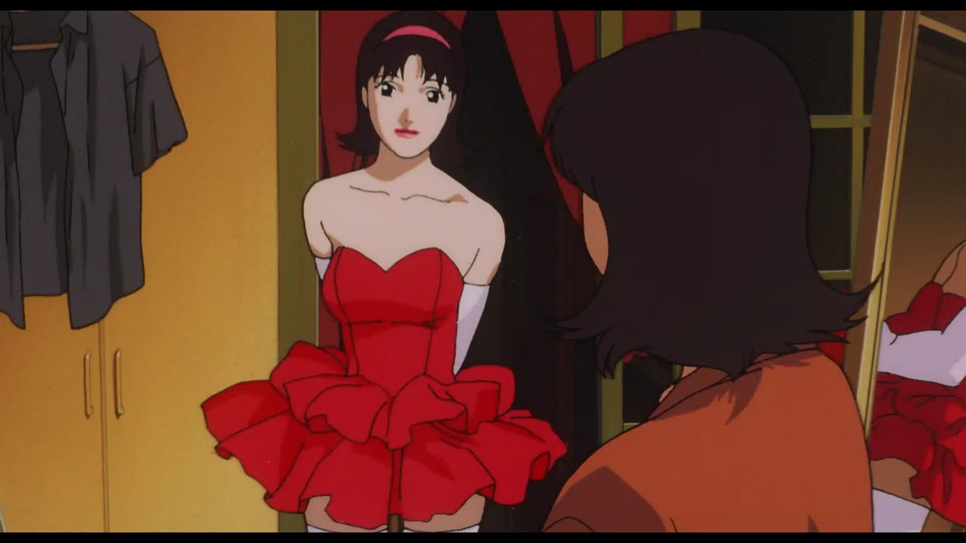 A woman in a frilly red dress stands in front of a mirror, looking at her reflection while another woman, seen from behind, faces her. A wardrobe with shirts is visible in the background.