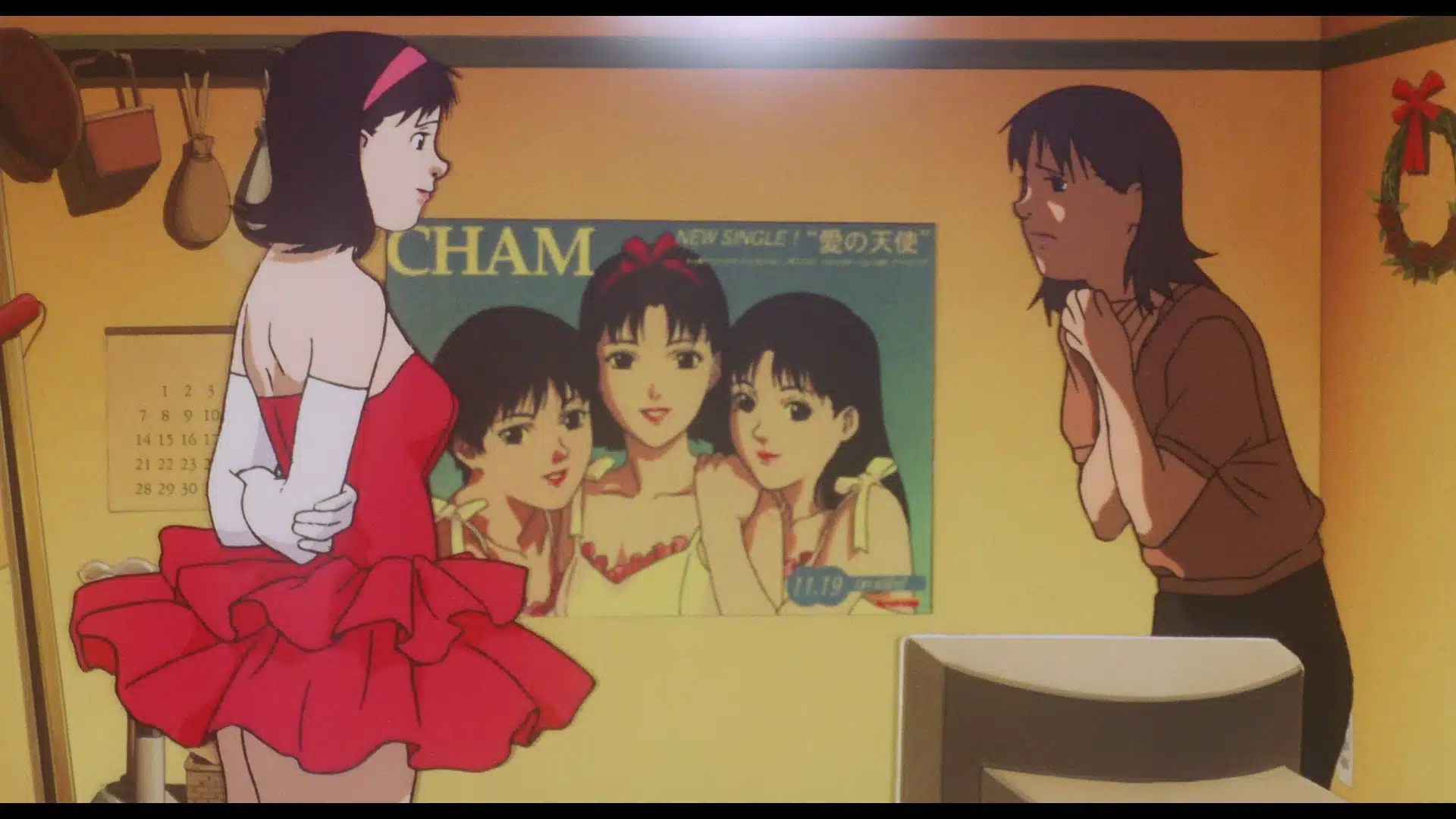 Two women stand in a room; one wears a red dress and white gloves, the other looks concerned. Behind them is a poster of three girls and a calendar on a yellow wall.