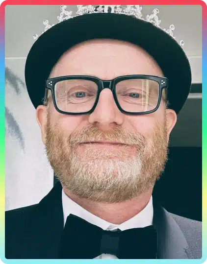 A person with a grey beard, wearing large black glasses, a black hat with a tiara, and a black dinner jacket with a bow tie, smiles at the camera. A colourful border frames the image, capturing the festive spirit of the Kilkenny Animated Festival.