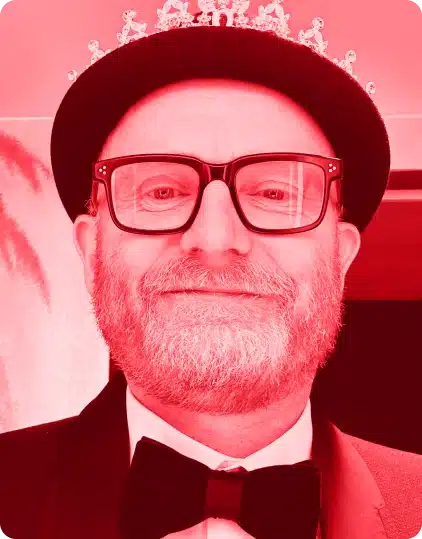 A bearded man wearing glasses, a bow tie, and a crown smiles at the camera during the Kilkenny Animated Festival. The image has a strong red tint.