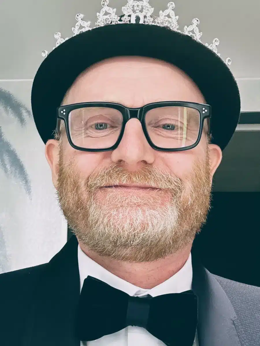 A bearded person in glasses, a black bowler hat, and dinner jacket smiles at the camera. They are also wearing a sparkly tiara over the hat.