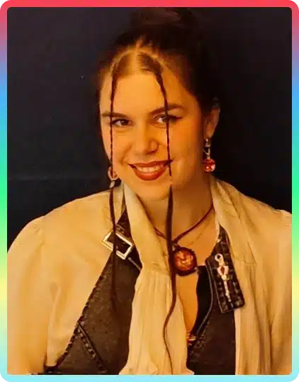 A smiling woman with dark hair in two front plaits wears earrings, a pendant necklace, and a layered outfit with a patterned shirt and cream scarf. The image has a rainbow border, capturing the vibrant spirit of the Kilkenny Animated Festival.