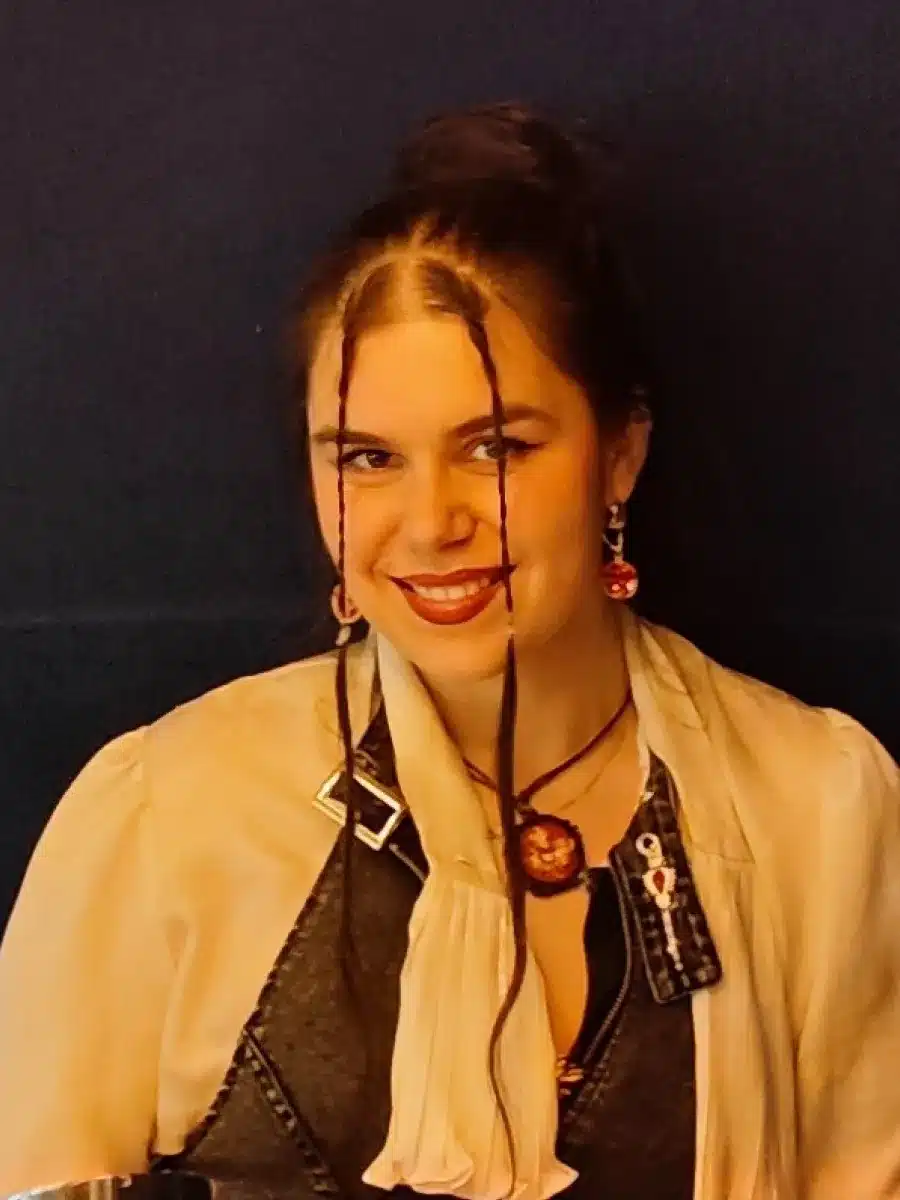 A woman with long brown hair and two thin plaits framing her face, wearing a light blouse, dark waistcoat, and statement jewellery, smiles in front of a dark background.