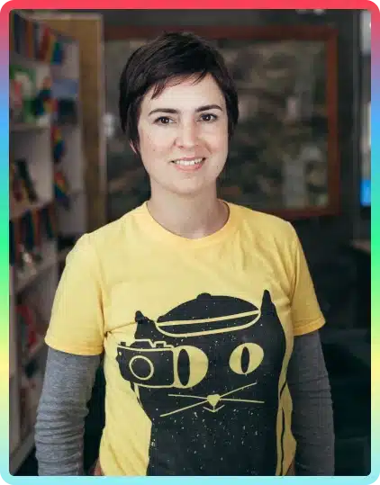 A person with short brown hair smiles, wearing a yellow T-shirt with a black cartoon cat design and a grey long-sleeve shirt underneath—perfect attire for the creative spirit of the Kilkenny Animated Festival. Shelves and a blurred background are visible.
