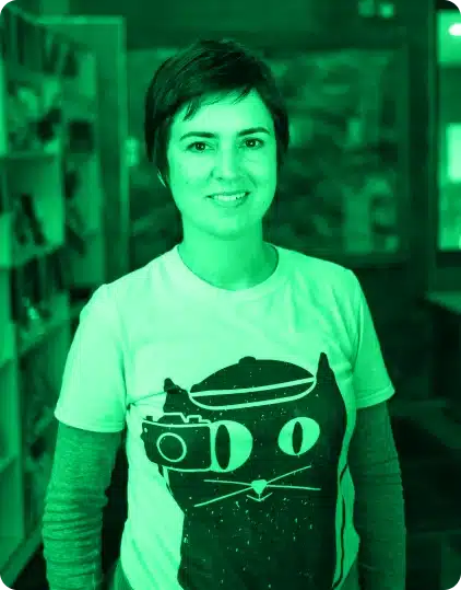 A person with short hair smiles at the camera, wearing a T-shirt featuring a cartoon cat with a camera and beret—reminiscent of the playful spirit of the Kilkenny Animated Festival. The image has a green filter. Shelves and a desk are visible in the blurred background.