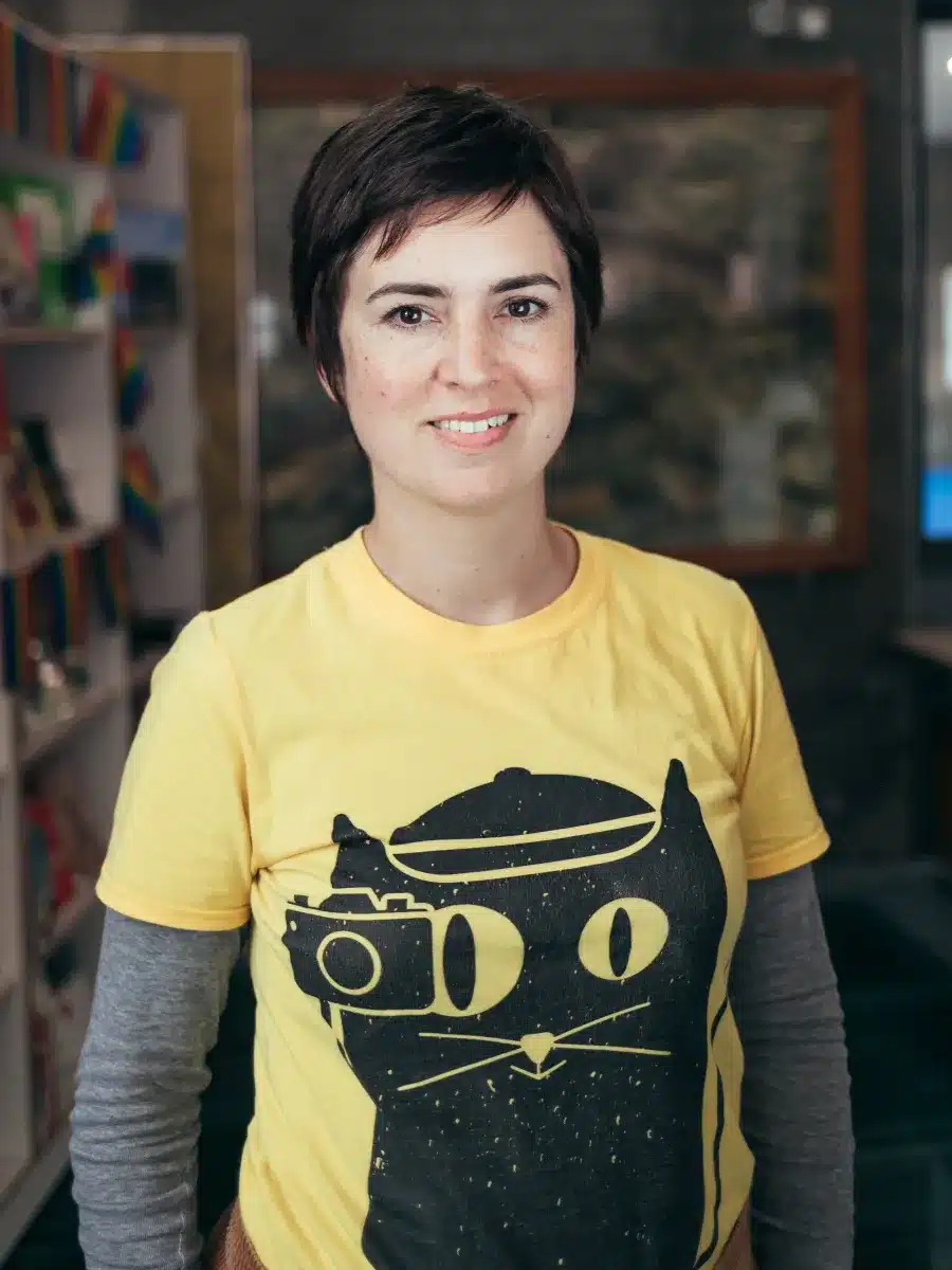 A smiling person with short brown hair wears a yellow shirt featuring a black cat graphic and stands indoors, with bookshelves and colourful items blurred in the background.