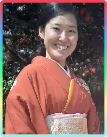 A woman wearing an orange kimono with floral patterns smiles outdoors in front of leafy trees and red flowers, capturing the vibrant spirit often seen at the Kilkenny Animated Festival.
