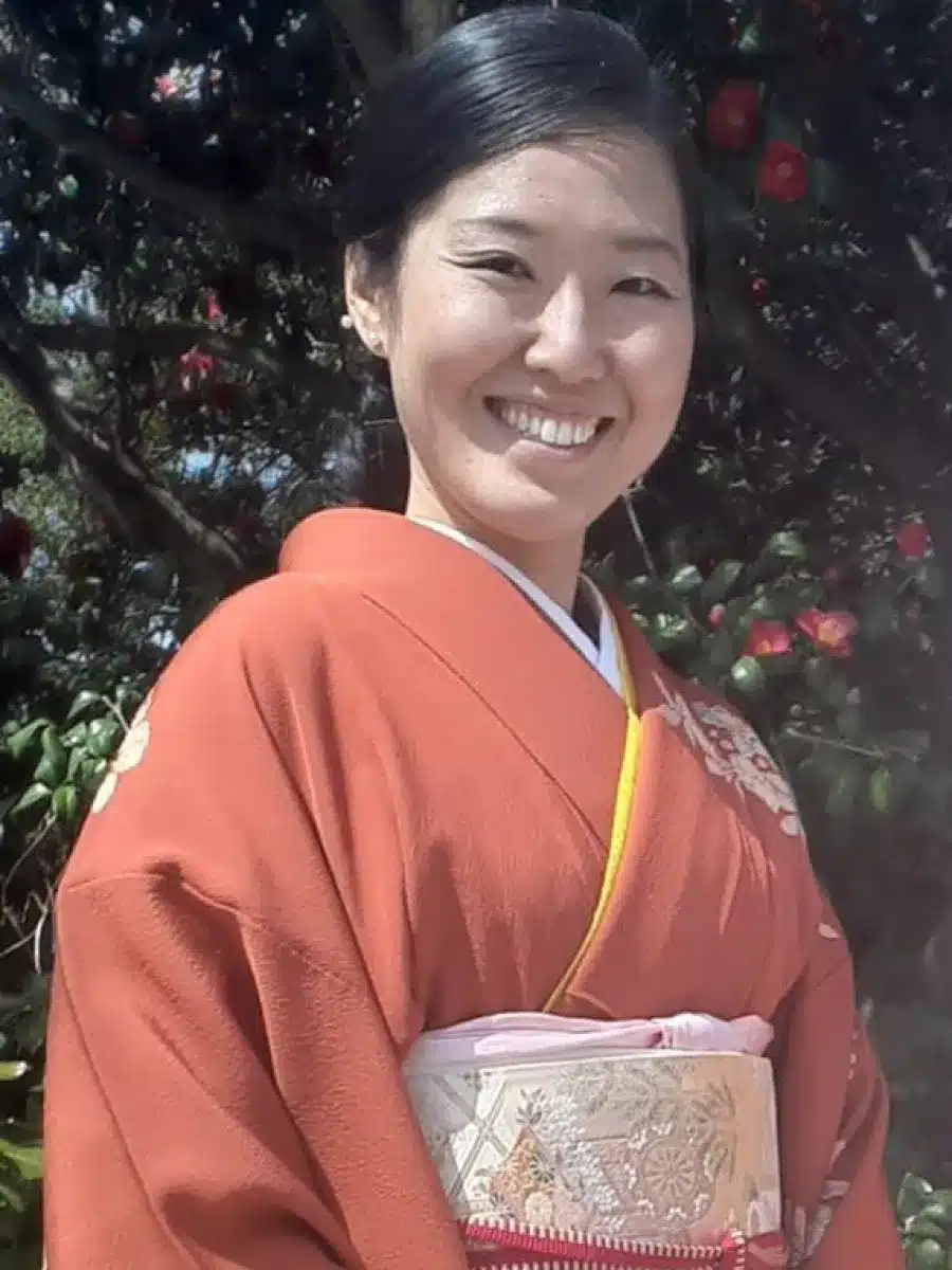 A woman smiles outdoors, wearing an orange kimono with floral patterns and a light-coloured obi. There are green leaves and red flowers in the background.